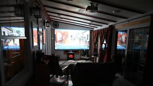 Een gedempt verlicht interieur met een groot televisiescherm waarop een video draait, een tafeltje met een stoel, en een open haard.