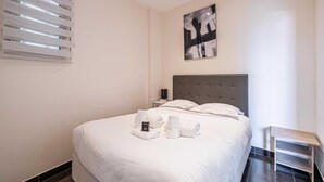 Een net opgemaakt bed met witte lakens, een nachtkastje en een schilderij aan de muur.