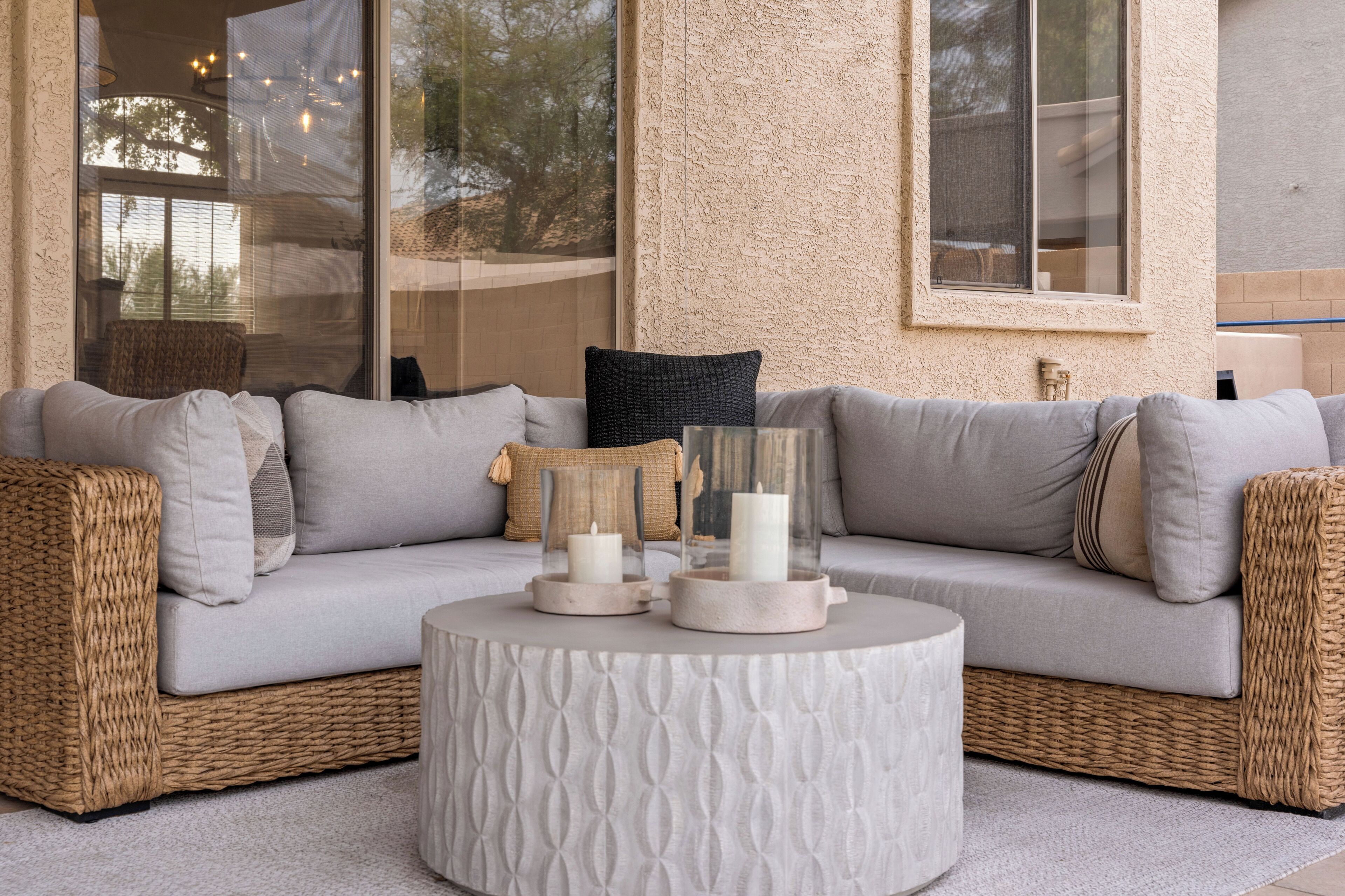 A wicker sofa with cushions, a round table with candles, and a window with a view of trees.