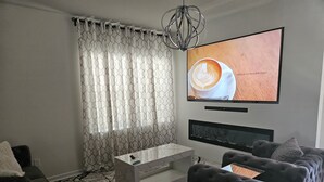 A modern living room with a large flat-screen TV mounted on the wall, a glass coffee table, a tufted sofa, and a patterned curtain. - Family-Friendly 3BR Fast WiFi  10 Min to Uptown (Charlotte)