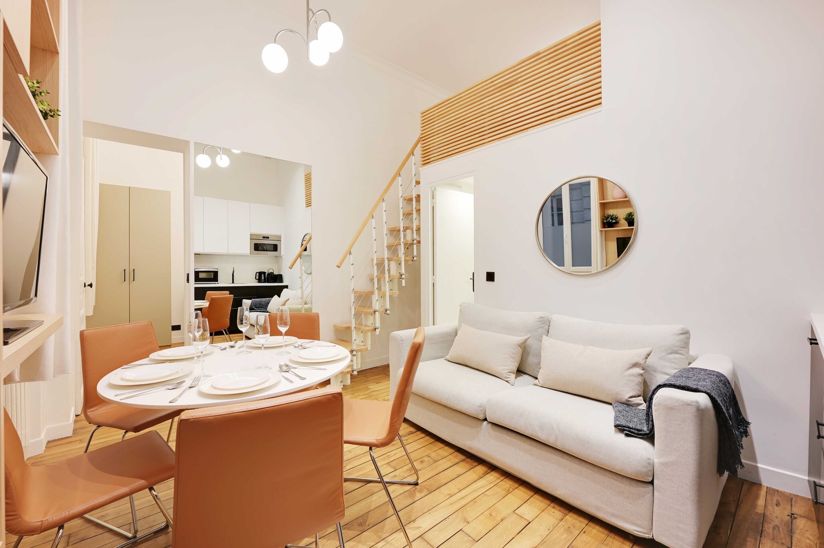 Un salon moderne avec un canapé blanc, une table à manger dressée pour quatre personnes et un escalier menant à un niveau supérieur.