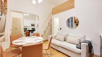 Un salon moderne avec un canapé blanc, une table à manger dressée pour quatre personnes et un escalier menant à un niveau supérieur.