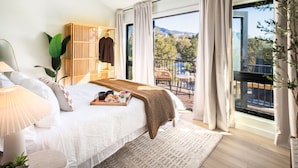 A bedroom with a large bed, a wooden wardrobe, a balcony with a table and chairs, and a view of trees and mountains. - A-Frame, HotTub, FirePit, Pergola, UFO Playscape, MiniAFrame ADU (Crestone)