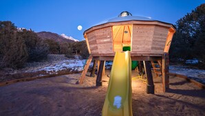 A wooden dome structure with a slide in the foreground, set in a natural landscape with mountains in the background. - A-Frame, HotTub, FirePit, Pergola, UFO Playscape, MiniAFrame ADU (Crestone)