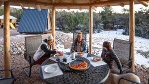 A covered outdoor dining area with a table set for three people, surrounded by natural scenery and a wooden structure in the background. - A-Frame, HotTub, FirePit, Pergola, UFO Playscape, MiniAFrame ADU (Crestone)