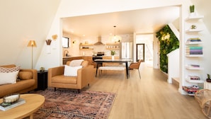 A spacious living area with a sofa, coffee table, and a patterned rug. A kitchen is visible in the background with a dining table and chairs. - A-Frame, HotTub, FirePit, Pergola, UFO Playscape, MiniAFrame ADU (Crestone)