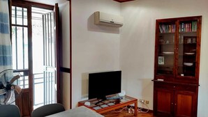 A living room with a television, a fan, a glass table, and a wooden cabinet with books.