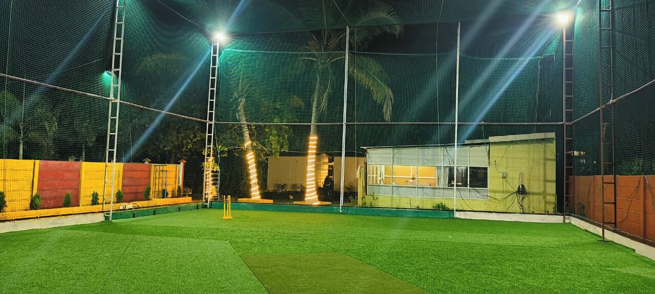 A well-lit cricket pitch with a netted enclosure, surrounded by a low fence and a small building in the background.