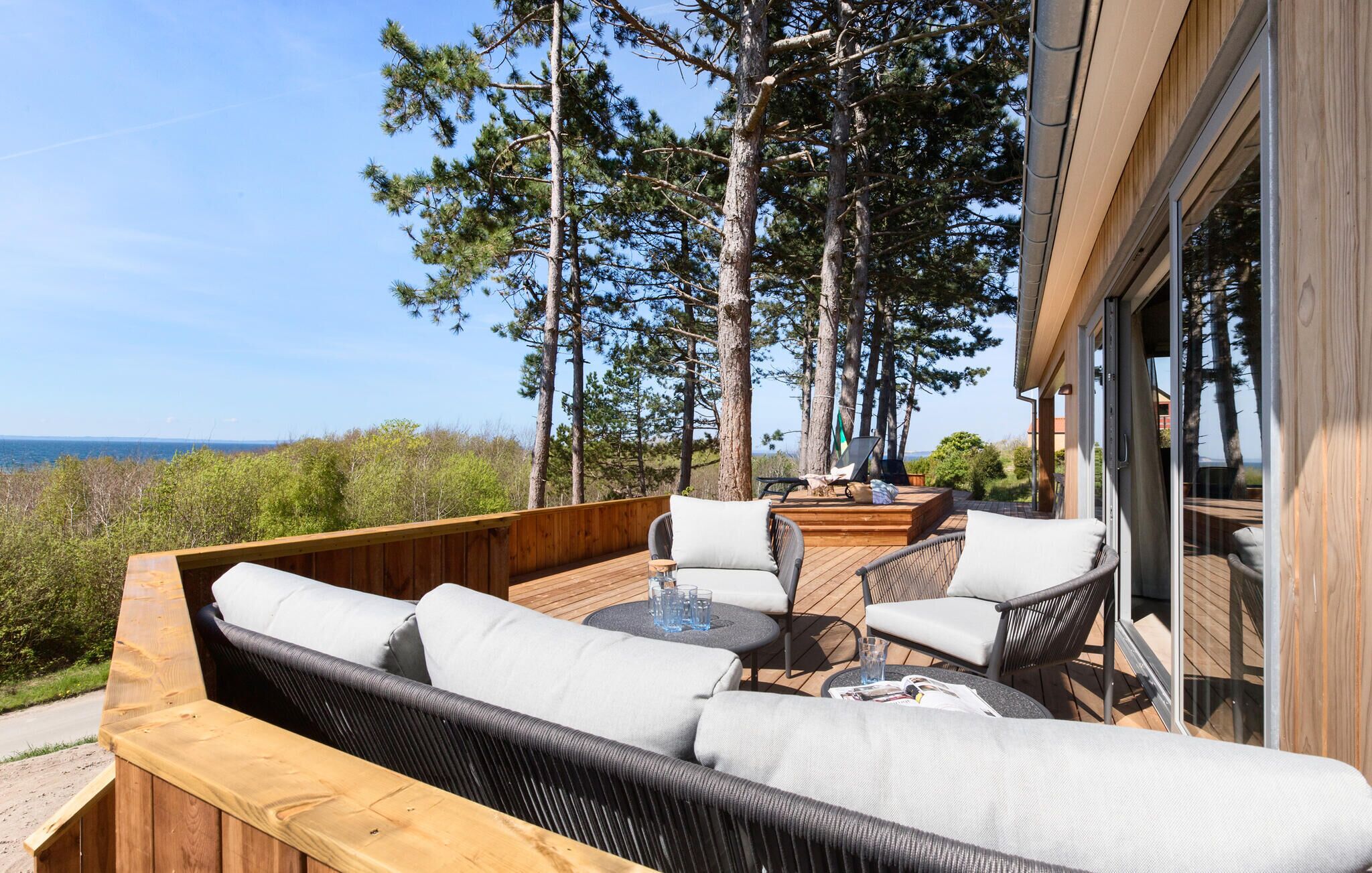 Eine Terrass mit Korbmöbeln, einem Glastisch und Blick auf das Meer und Bäume.