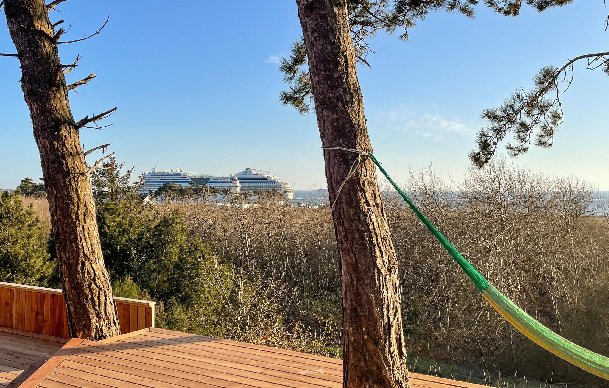 A wooden deck with a hammock, trees, and a view of cruise ships in the distance.
