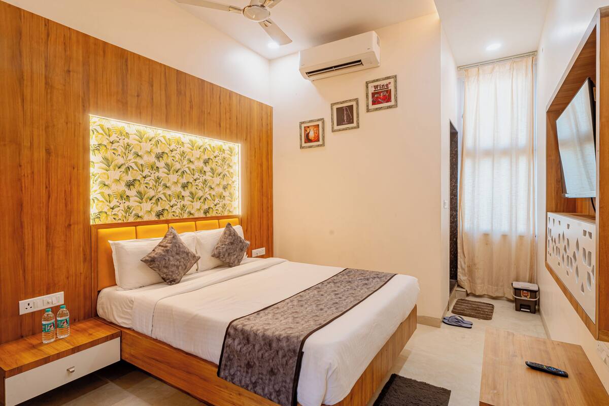 A hotel room with a wooden headboard, a bed with white linens, a bedside table with a lamp, and a wall-mounted TV.