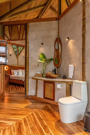 A room with a wooden floor, a bed, a bathroom with a toilet, a sink, and a mirror.