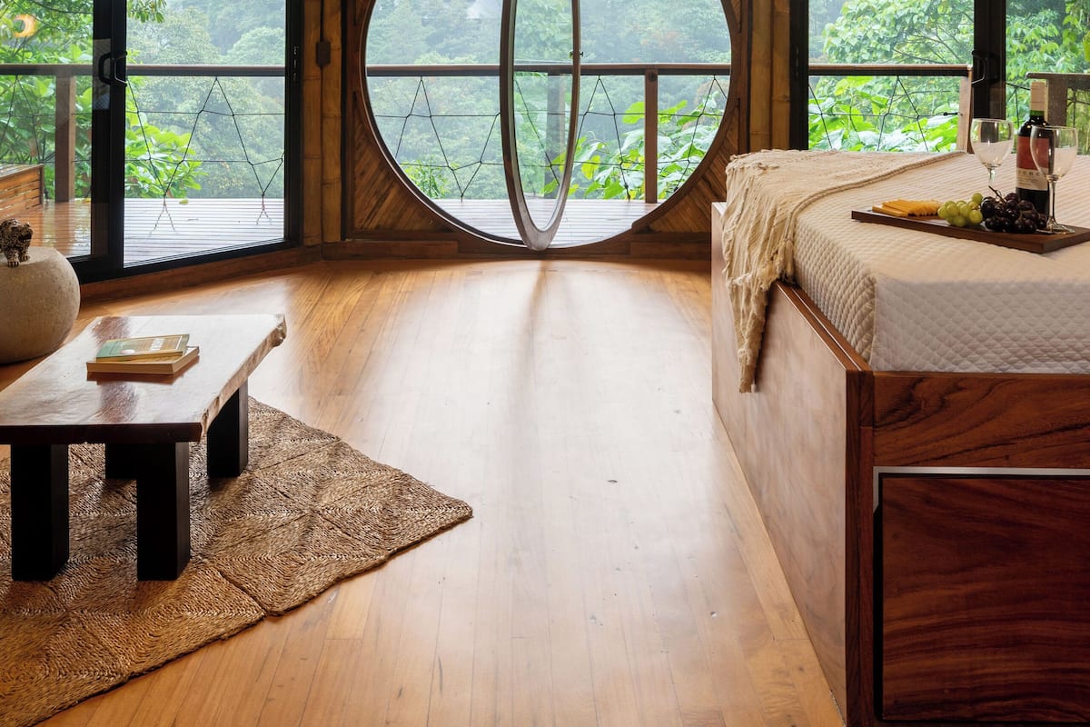Quarto com uma grande janela redonda, uma cama, uma mesa de madeira e um candeeiro suspenso.