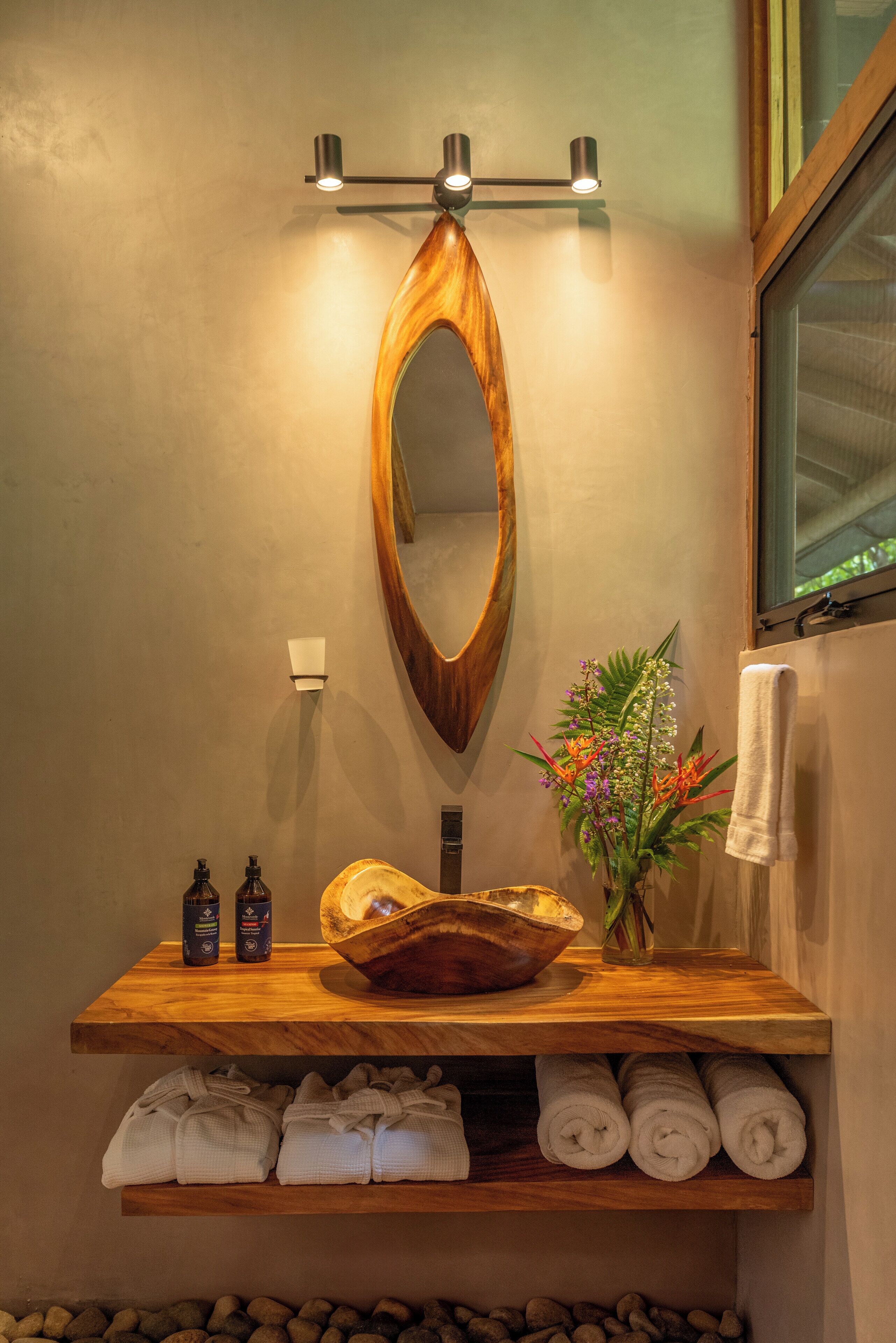 A bathroom with a wooden vanity, a large mirror, a bowl sink, a vase with flowers, and towels neatly stacked below the vanity.