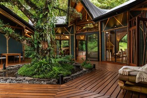 Une terrasse en bois agrémentée d’un grand arbre et d’une végétation luxuriante, entourée de portes vitrées donnant sur un espace repas.