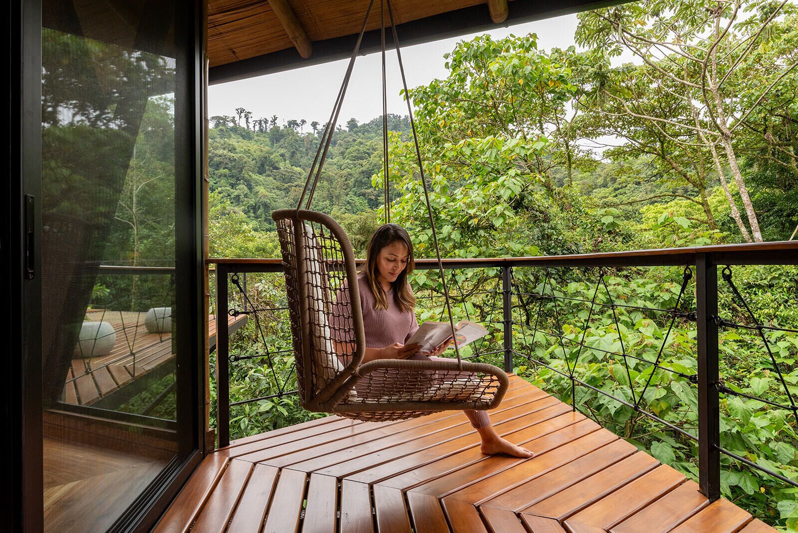A person sitting on a wicker swing chair on a wooden deck with a view of lush greenery.