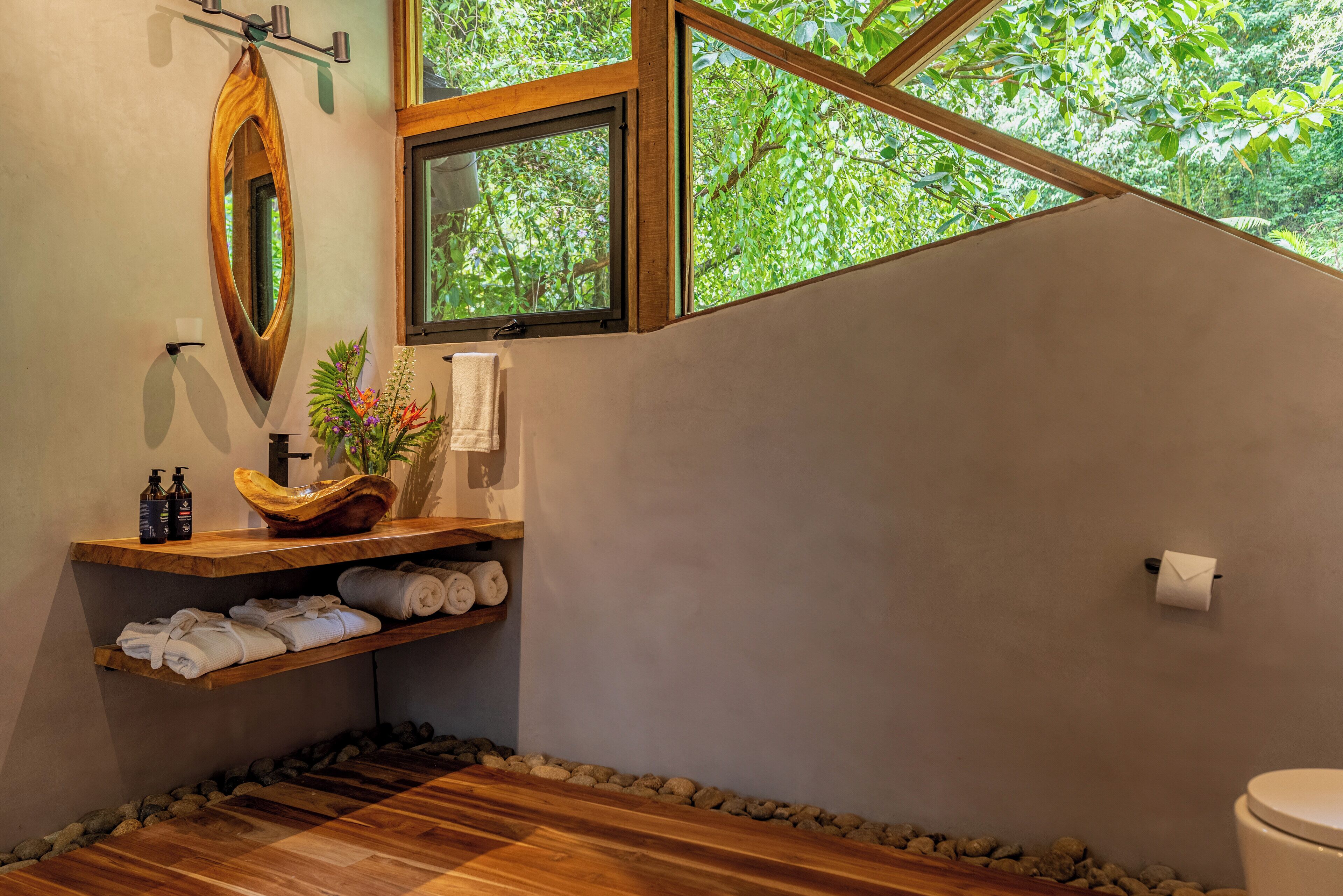 A bathroom with a wooden vanity, a large mirror, a window with a view of trees, and a toilet.