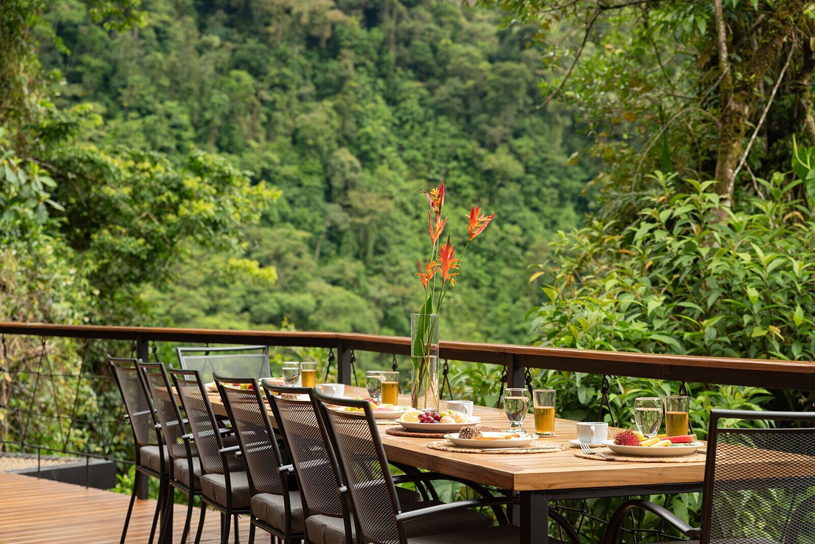 Un espacio de comedor al aire libre con una mesa y sillas de madera, rodeado de abundante follaje y un paisaje boscoso.