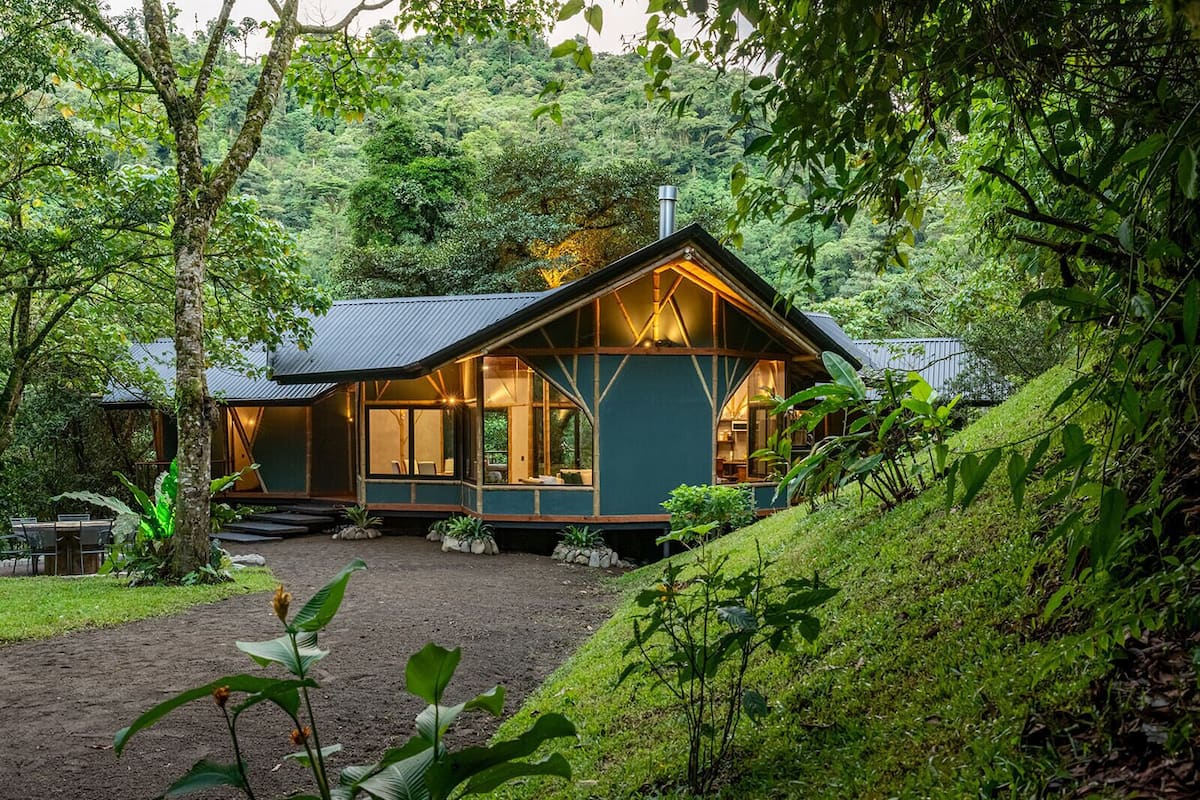 Uma cabana rústica com telhado inclinado, rodeada por vegetação exuberante e um caminho de terra que leva à entrada.