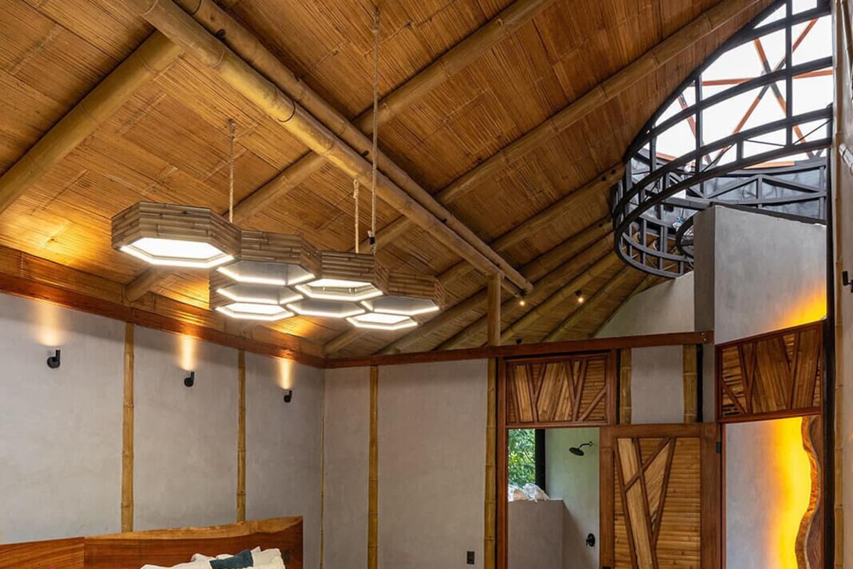 Quarto de hotel moderno com teto de madeira, uma cama grande, uma cama adicional, um espaço de estar com uma cadeira e uma mesa pequena, e vista para o exterior pela janela.