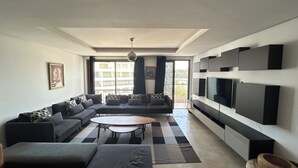 A modern living room with a sectional sofa, a round coffee table, and a flat-screen TV mounted on the wall. - THE ESCAPE FROM BOUREGREG (Salé)