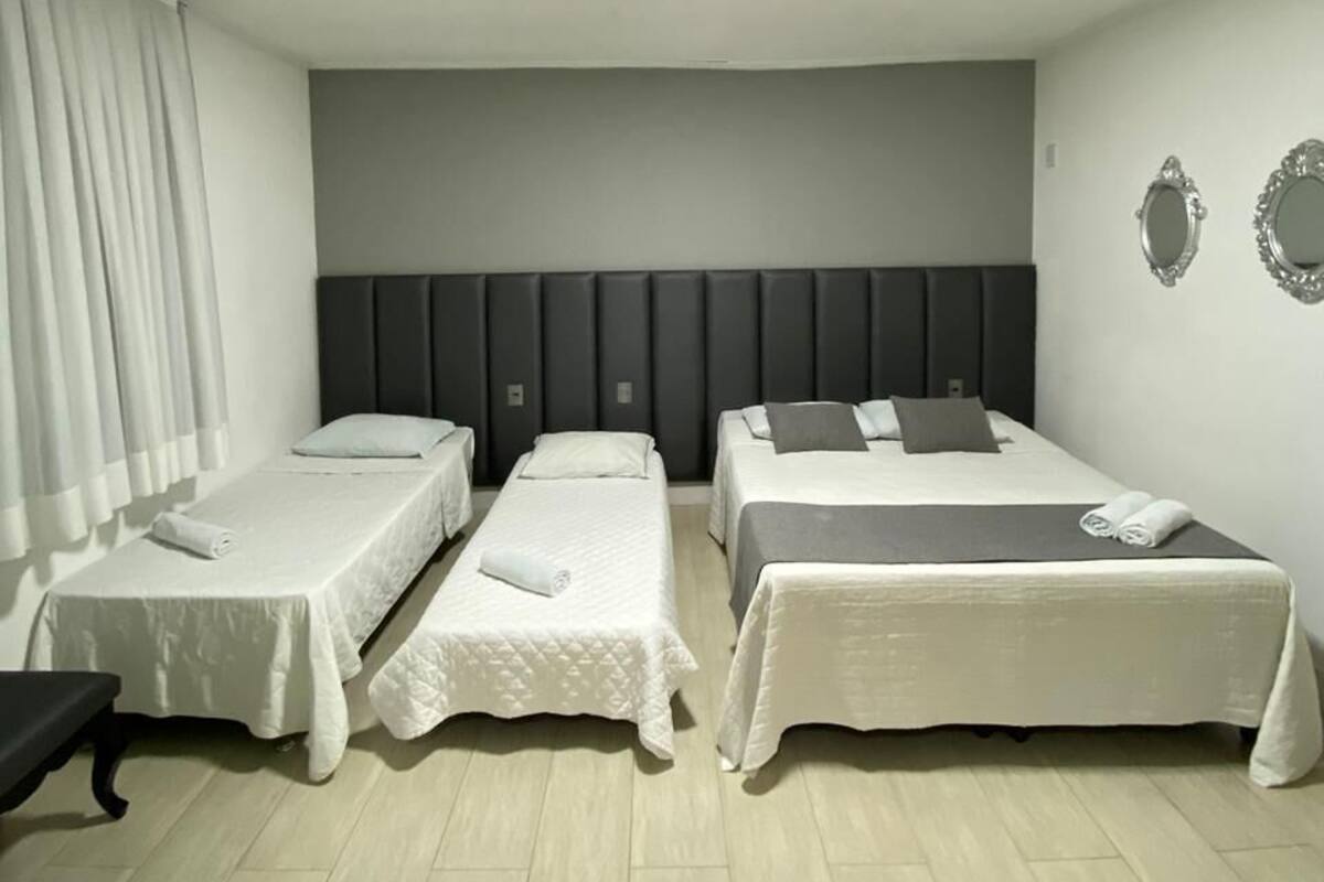 Quarto de hotel com duas camas de solteiro, uma cama grande, um banco e um espelho na parede.