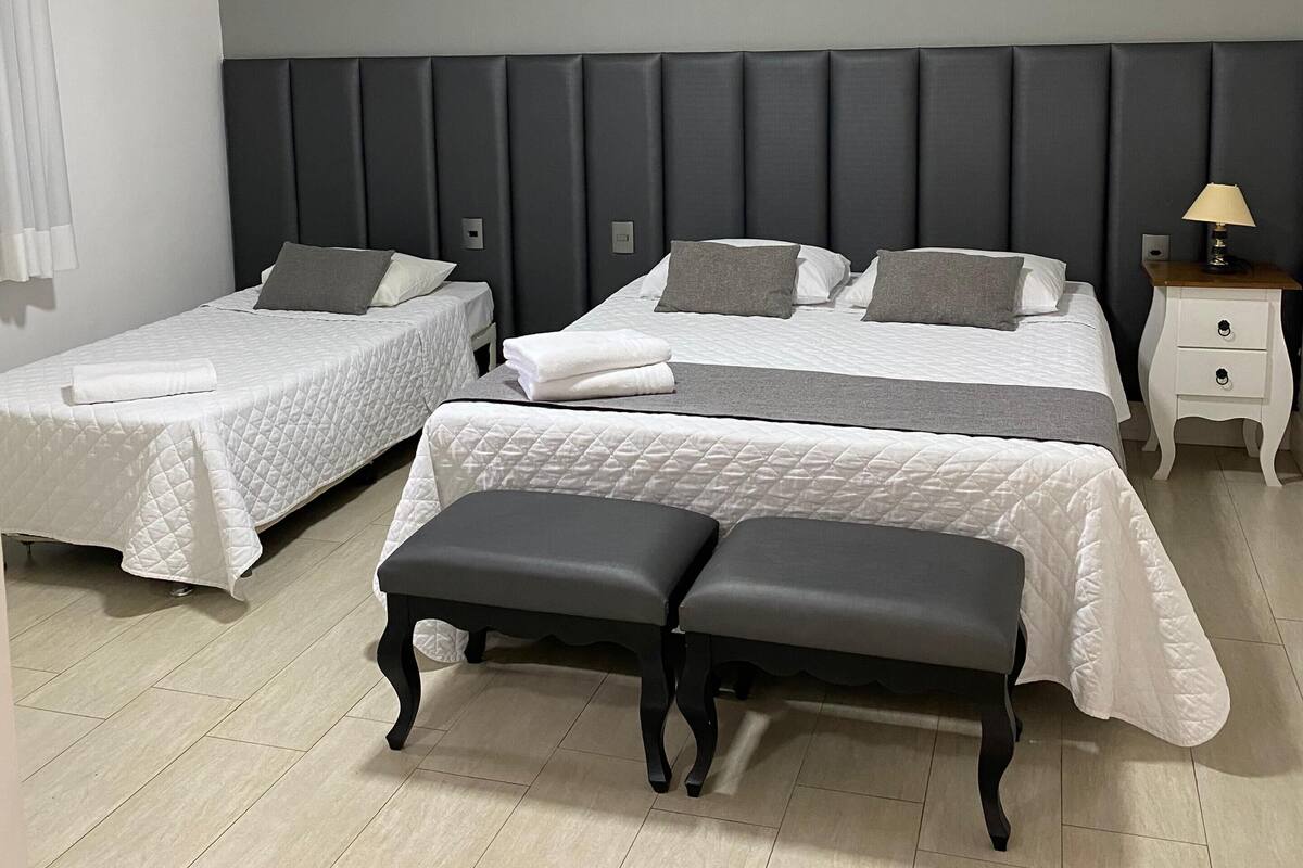 Quarto de hotel com duas camas de solteiro, uma cama de casal, um banco, uma mesinha de cabeceira com abajur e uma luminária no teto.