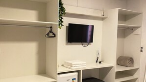 A room with a white desk, a flat-screen TV mounted on the wall, a small refrigerator, and a bench. - The Reds Pousada (Penha)