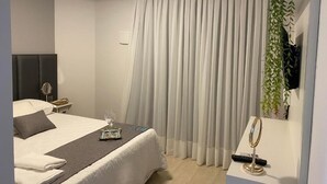 A modern hotel room with a neatly made bed, a bedside table with a lamp, a large window with curtains, and a small plant on the wall. - The Reds Pousada (Penha)