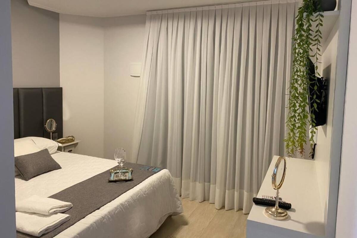 Quarto de hotel moderno com uma cama bem arrumada, mesa de cabeceira com luminária, uma grande janela com cortinas e uma pequena planta na parede.