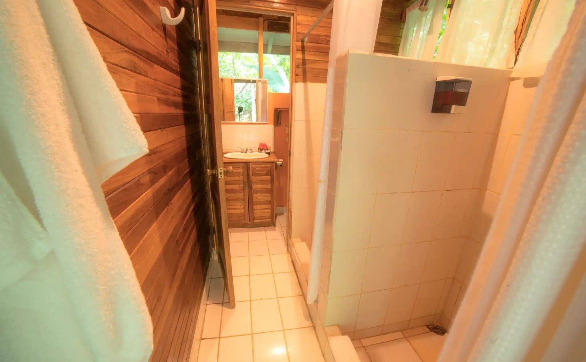 A narrow hallway with wooden walls and a tiled floor leading to a bathroom with a shower and a sink.