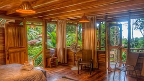 A wooden room with a bed, a chair, a fan, and large windows offering a view of lush greenery. - Samasati Rainforest Bungalows (Cahuita)