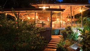 A wooden structure with a covered patio area, surrounded by lush greenery and a stone pathway leading to the entrance. - Samasati Rainforest Bungalows (Cahuita)