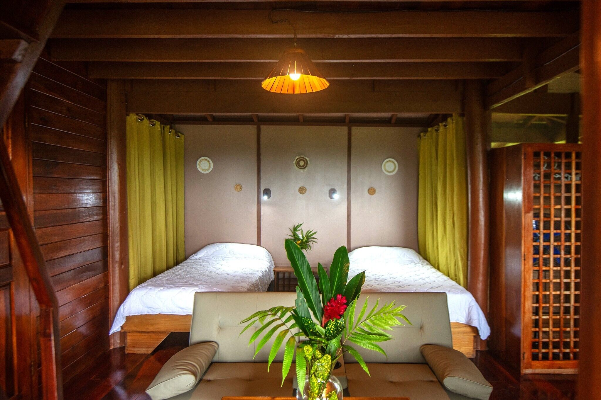 A room with two beds, a wooden ceiling, a hanging light fixture, and a small table with a vase of flowers.