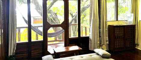 A spacious room with a large bed, a wooden table, and a view of the outdoors through large windows.