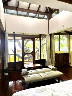 A spacious room with a large bed, a wooden table, and a view of the outdoors through large windows. - Samasati Rainforest Bungalows (Cahuita)