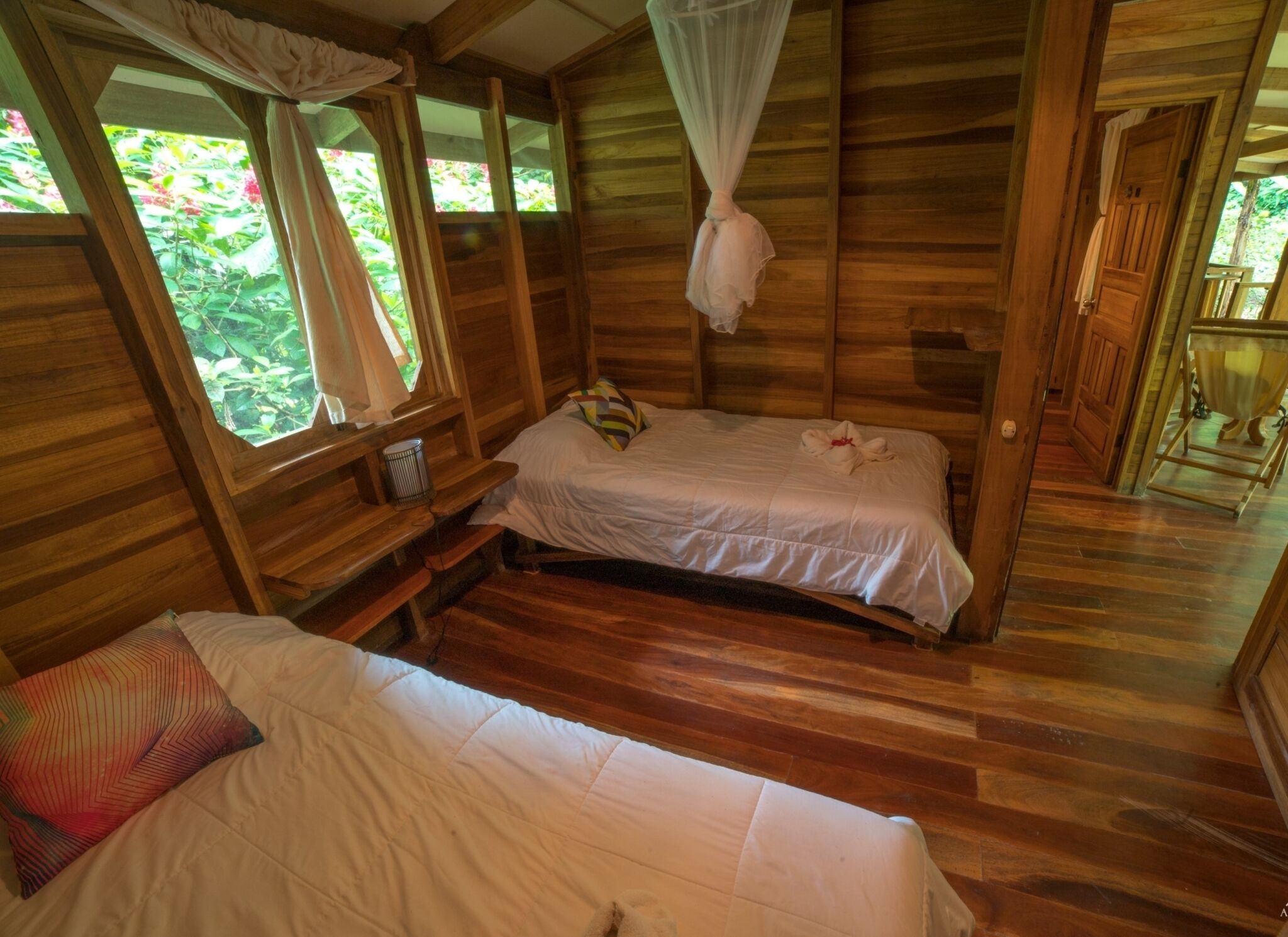 A wooden cabin with two beds, a small table, and a window with a view of greenery.