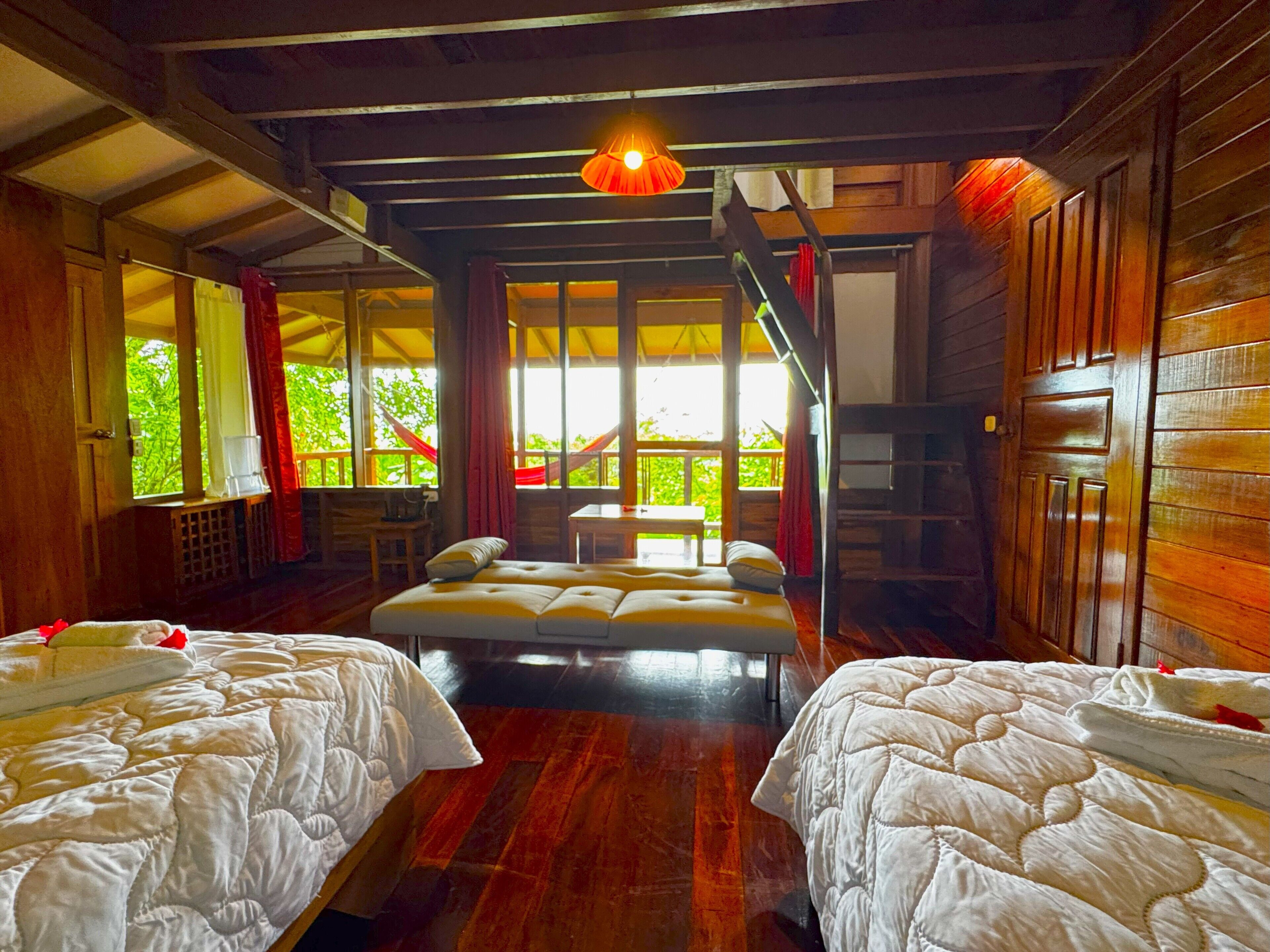 Une chambre avec deux lits, un plancher en bois, une grande fenêtre avec des rideaux et un luminaire suspendu.