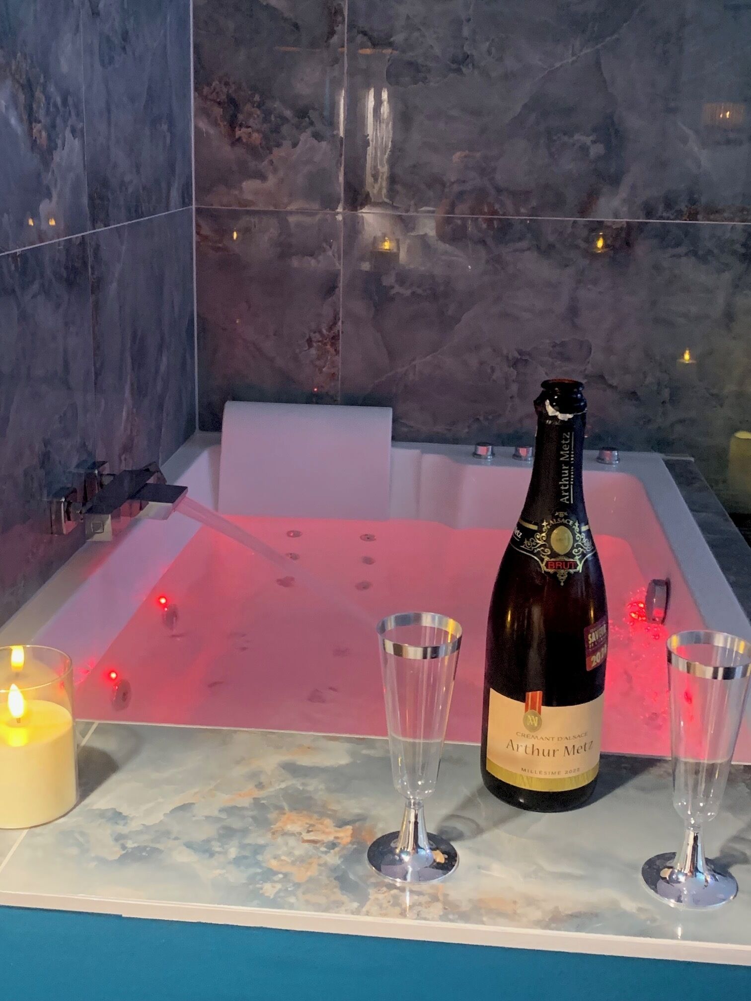 Una botella de champán Arthur Metz, dos copas y una vela encendida sobre una superficie de mármol en una habitación con una bañera y un muro de azulejos.
