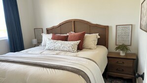 Ein ordentlich bezogenes Bett mit Holz-Kopfteil, mehreren Kissen, einem Nachttisch mit Lampe und einem kleinen Topfpflanzen.