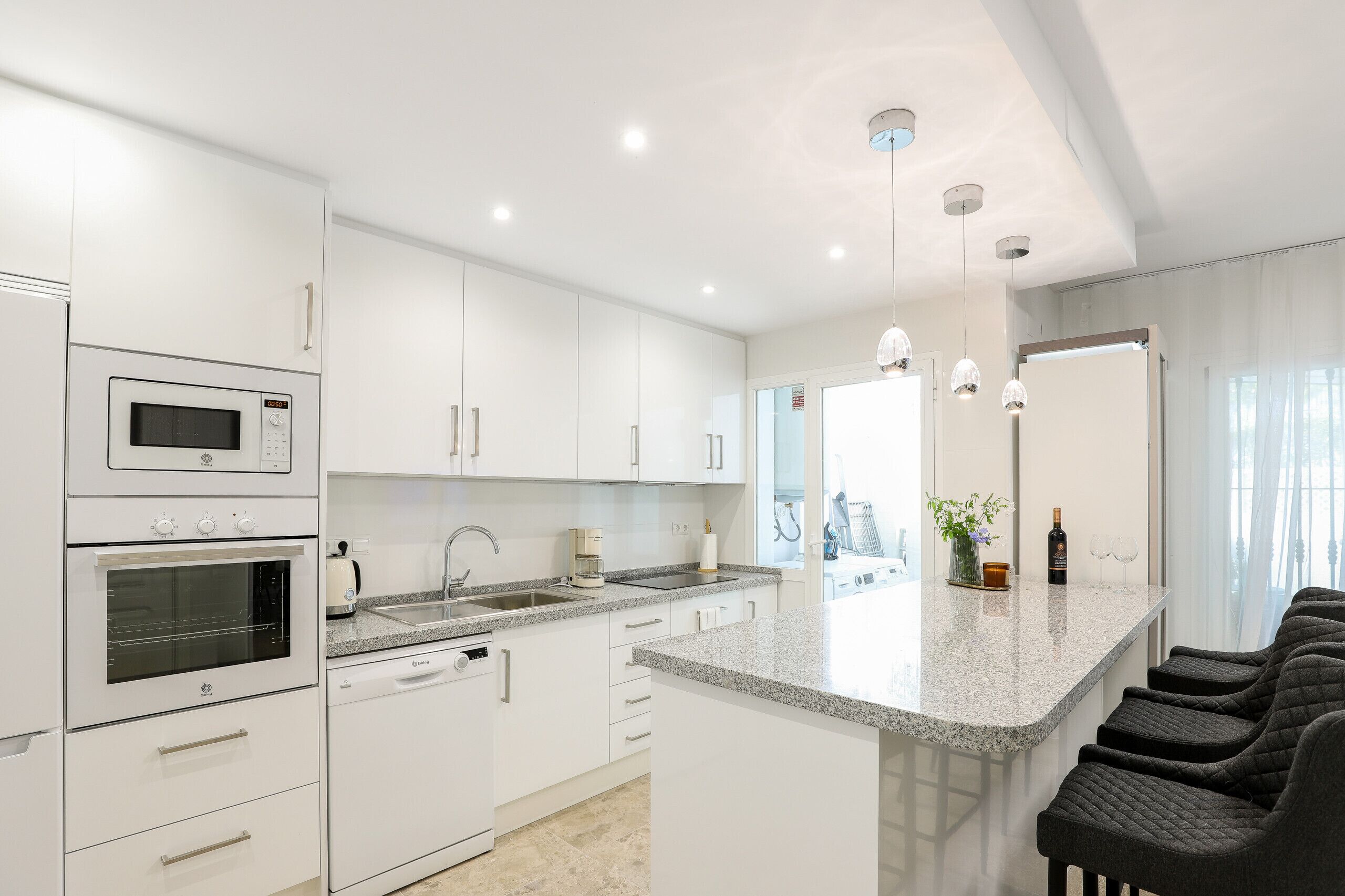 Una cocina moderna con armarios blancos, horno integrado, pileta y encimera de granito. Tres lámparas colgantes iluminan la isla.