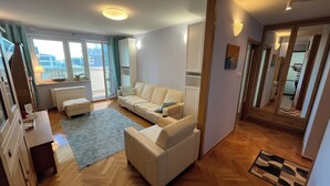 Ein modernes Wohnzimmer mit einer Couch, einem Couchtisch und einem Teppich. Durch das Fenster ist ein Ausblick, und eine Tür führt in einen anderen Raum.