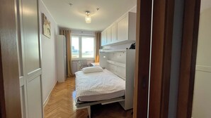 Ein schmaler Flur, der zu einem Schlafzimmer mit Bett, Heizkörper und einem Vorhangfenster führt.