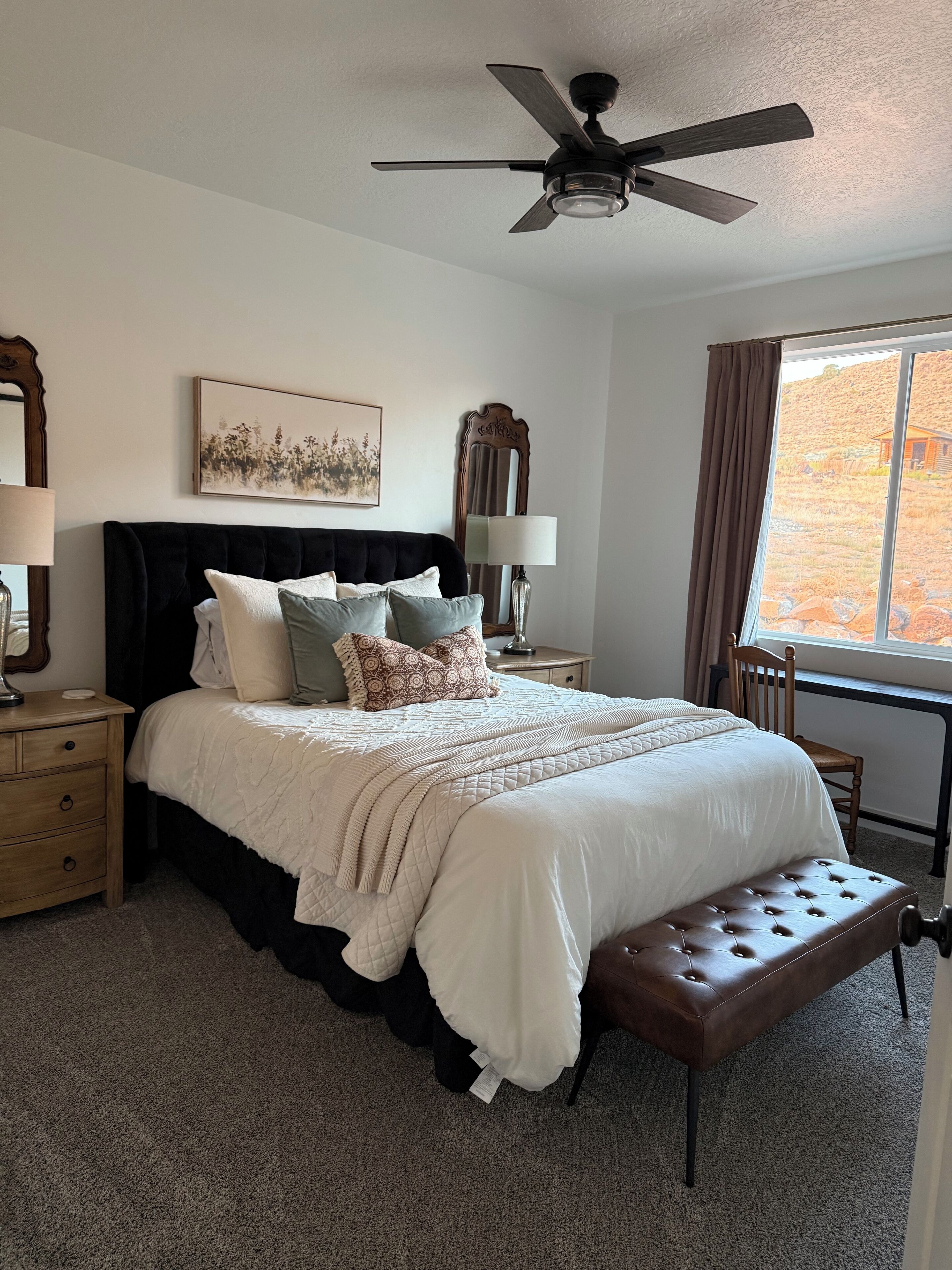 A bedroom with a large bed, a ceiling fan, a window with a view of a hilly landscape, a bedside table with a lamp, and a nightstand with a lamp.