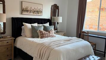 A bedroom with a large bed, a ceiling fan, a window with a view of a hilly landscape, a bedside table with a lamp, and a nightstand with a lamp.