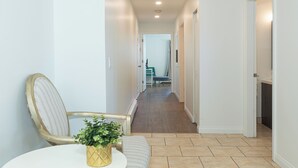A narrow hallway with a chair and a small table with a plant, leading to a bathroom with a shower and a chair.