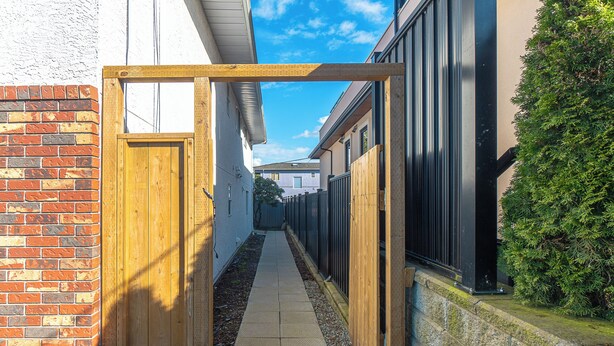 A narrow alleyway with a wooden gate, brick wall, and a pathway leading to a building with a black metal fence and a green bush.