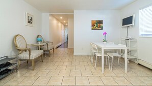 A dining area with a white table and chairs, a TV mounted on the wall, and a hallway leading to another room.