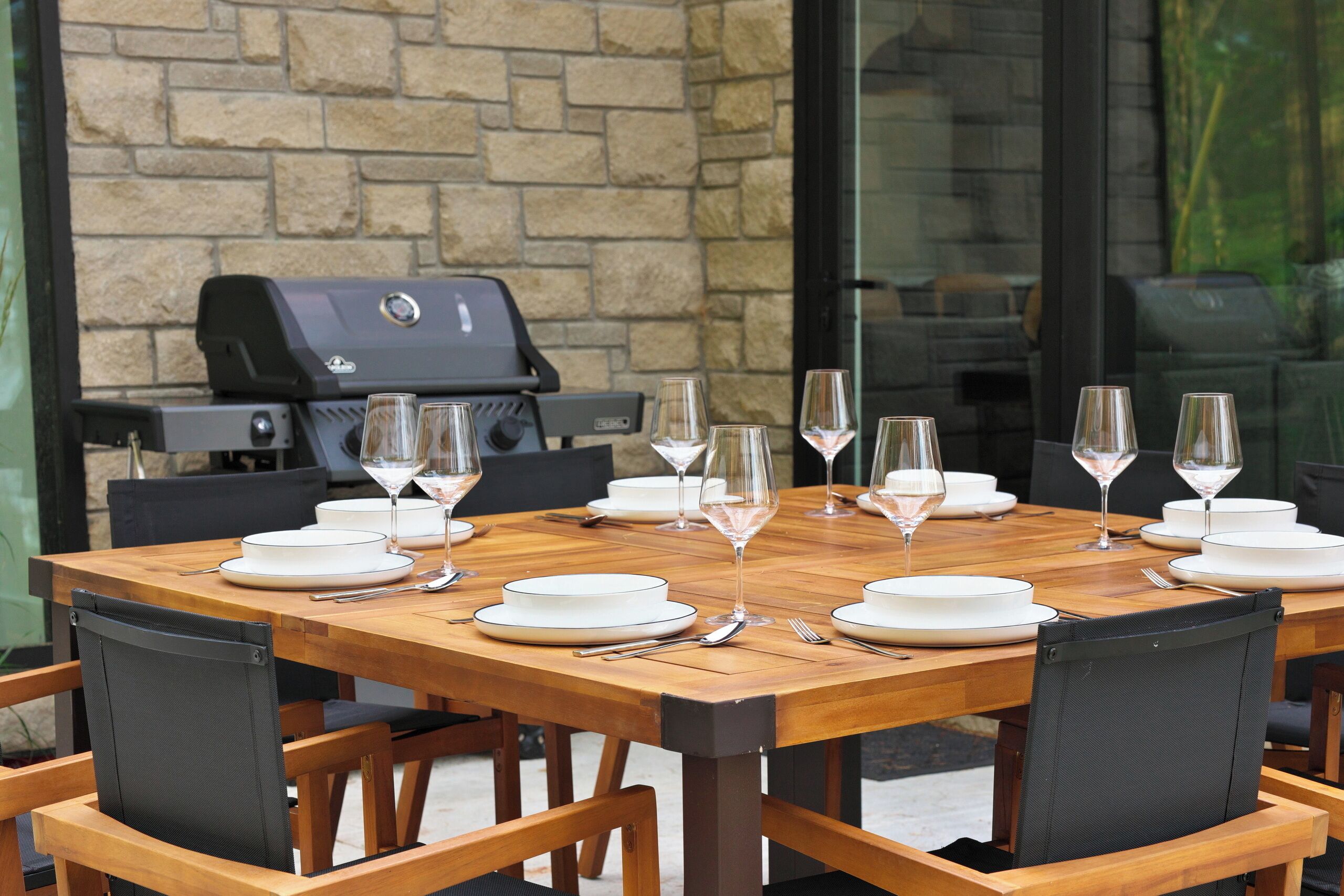 Una mesa de comedor redonda de madera dispuesta para seis personas, con platos blancos y copas de vino, una parrilla al fondo y una pared de piedra junto a una puerta de cristal.
