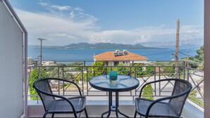 A balcony with two wicker chairs and a small round table, overlooking a coastal view with mountains and a house with a red roof. - Studio Apartment 'Two Adults No 1' with Sea View, Wi-Fi and Air Conditioning (Nikiana)